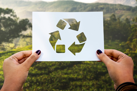 The recycling symbol is cut out on a piece of paper, held out in an outdoor setting.